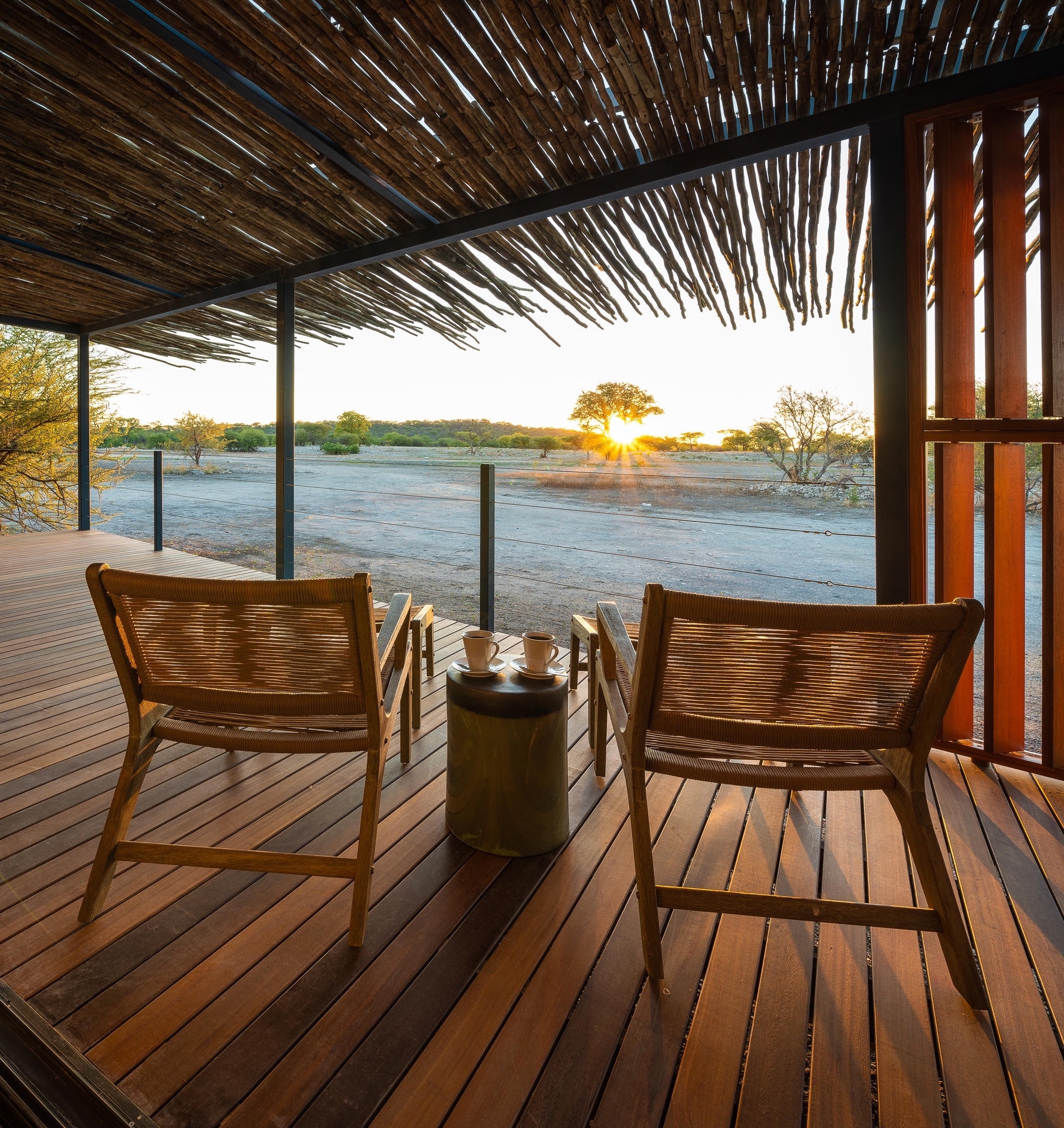 A serene wooden patio with two chairs and a small table overlooking a sunset in a peaceful landscape.
