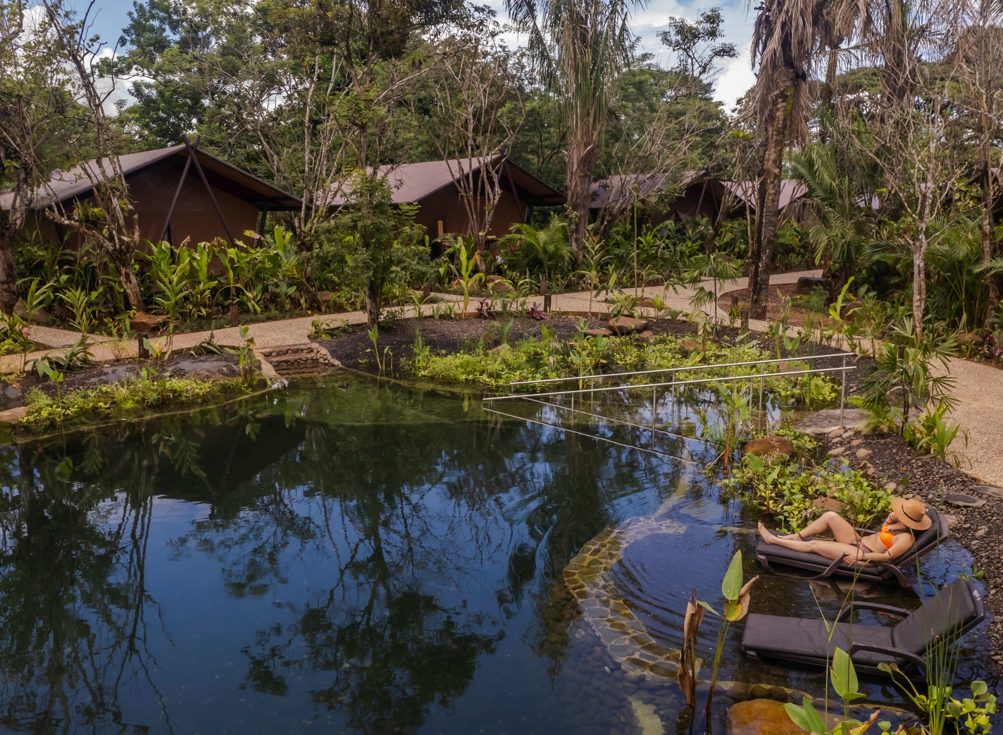 A person in a swimsuit sits on a dark grey lounge chair next to a small body of water surrounded by green plants, palm trees, jungle and in the background three low lodge bungalows are tucked into trees.