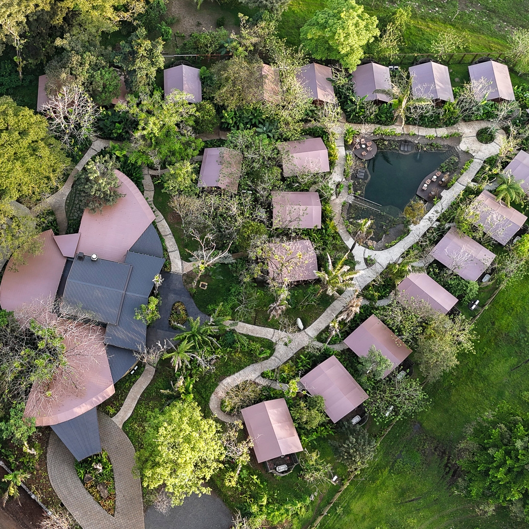 An aerial shot of a hotel with a main lodge building on the left and 18 smaller buildings can be seen to the right with some small walkways connecting them and lots of green trees and grass in between them.