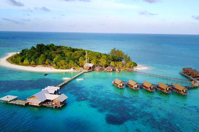Aerial shot of a resort on a tiny island surrounded by white sand and turquoise water and covered in dark palms. A long pier extends from one side with a building over the water, and on the other side, a row of smaller villas run perpendicular to the beach over the water.