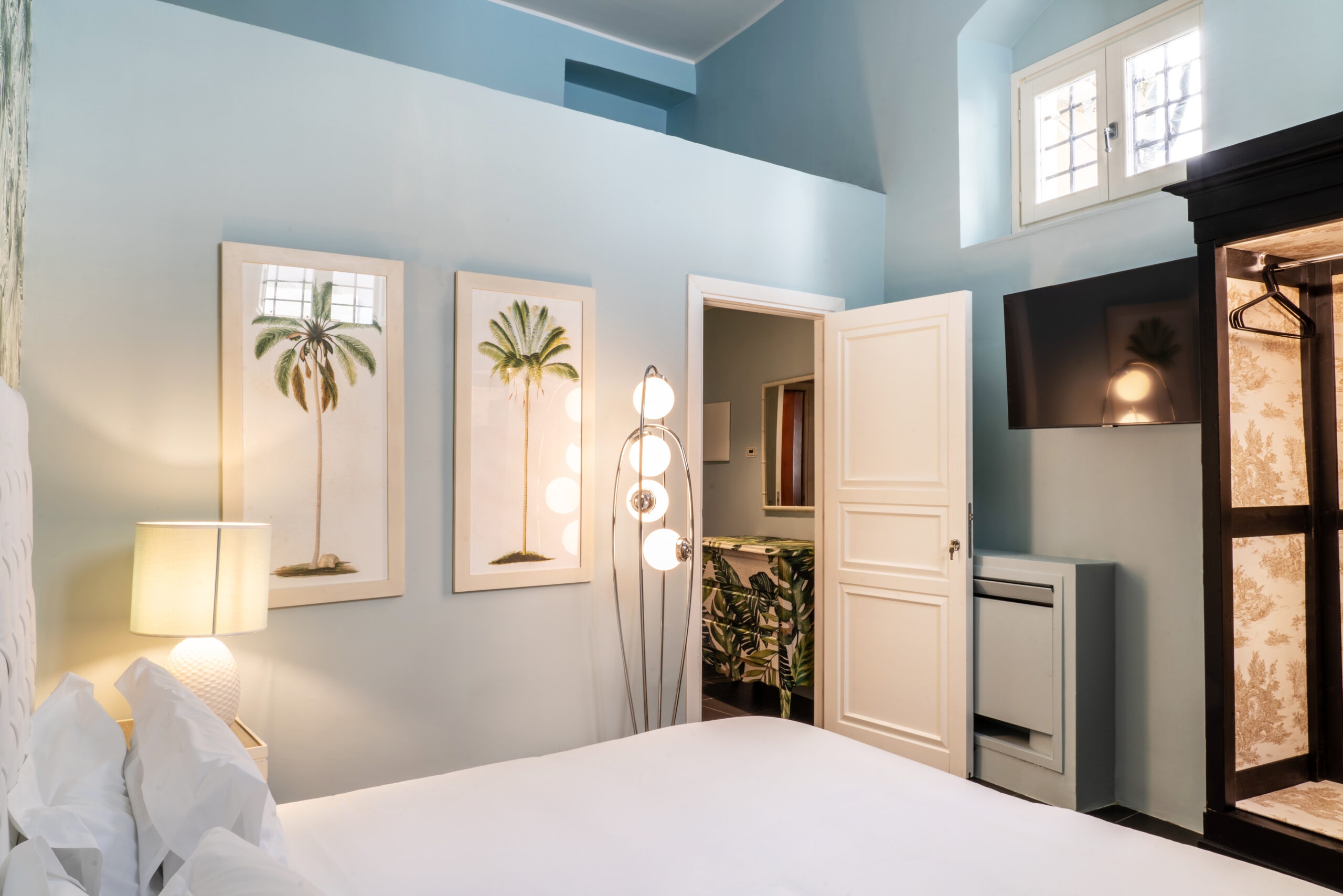 Bedroom with light blue walls, a bed with white sheets, an open bathroom door and on the wall two paintings of palm trees.