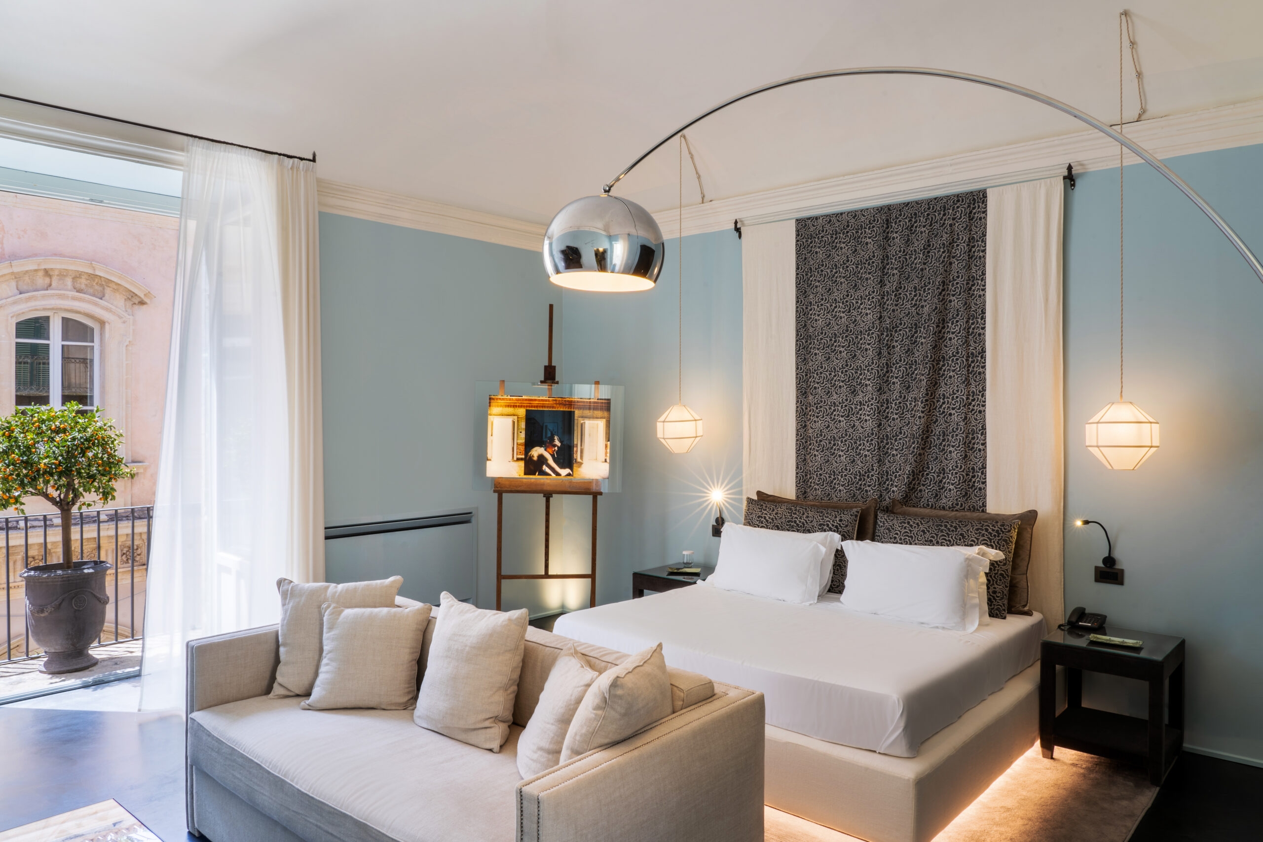 Interior shot of a bedroom with a double bed with grey sheets and a sofa at the foot of the bed with grey pillows. A large silver lamp looms over the bed. The walls are light blue and there is a window opening onto a julliette balcony with a decorative tree.
