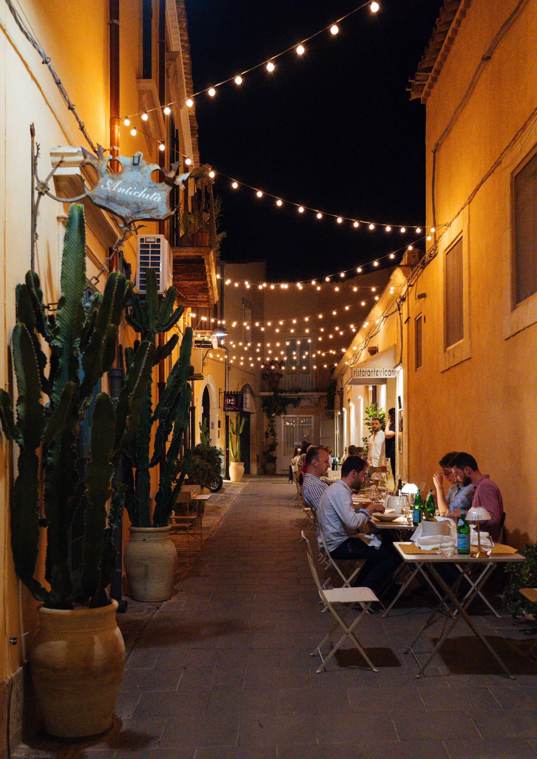 Exterior of a pedestrian street in the evening with a few tables where people are seated, and overhead are strung fairy lights.