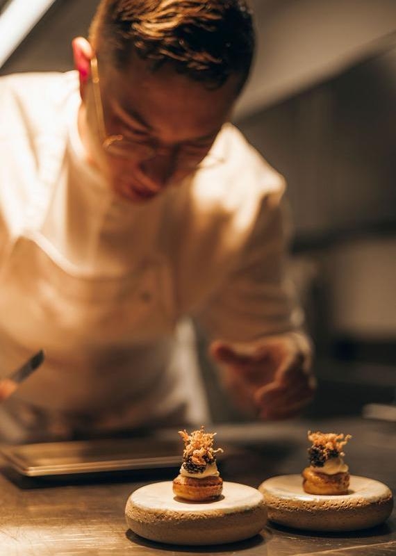 Close-up of Chef Davor Arnautović plating two delicate gourmet pastries on stone pedestals in a warm kitchen.