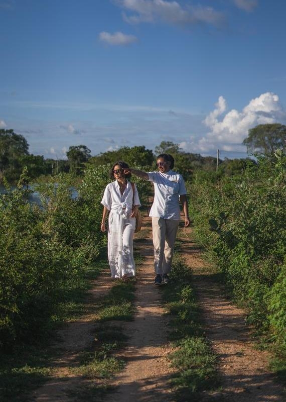 Two people in white outfits walking along a dirt trail through green bushes under a blue sky.