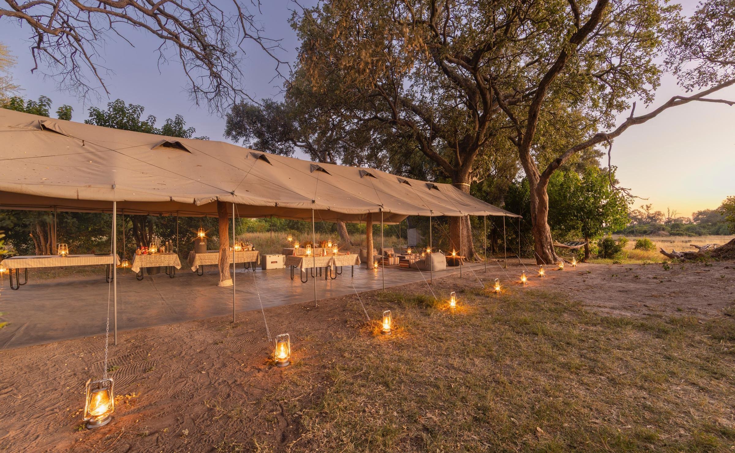 A large tan safari mess tent illuminated by a row of lanterns on the ground during a golden sunset.