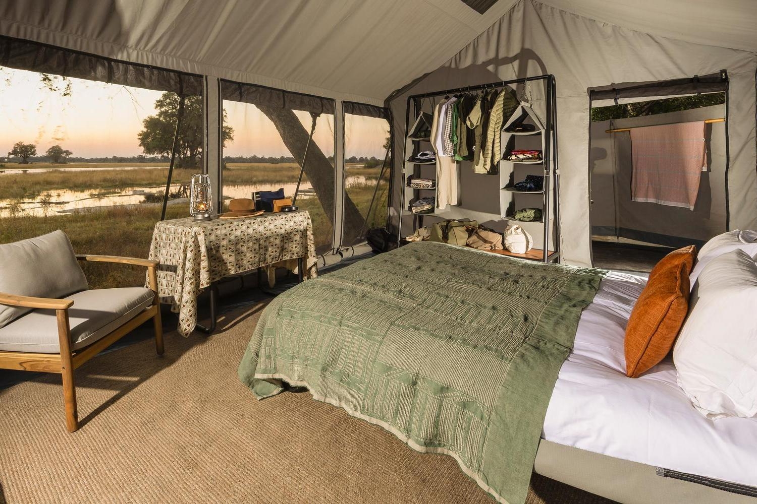 Interior of a safari tent with a green bedspread, a small table with a lantern, and large mesh windows.