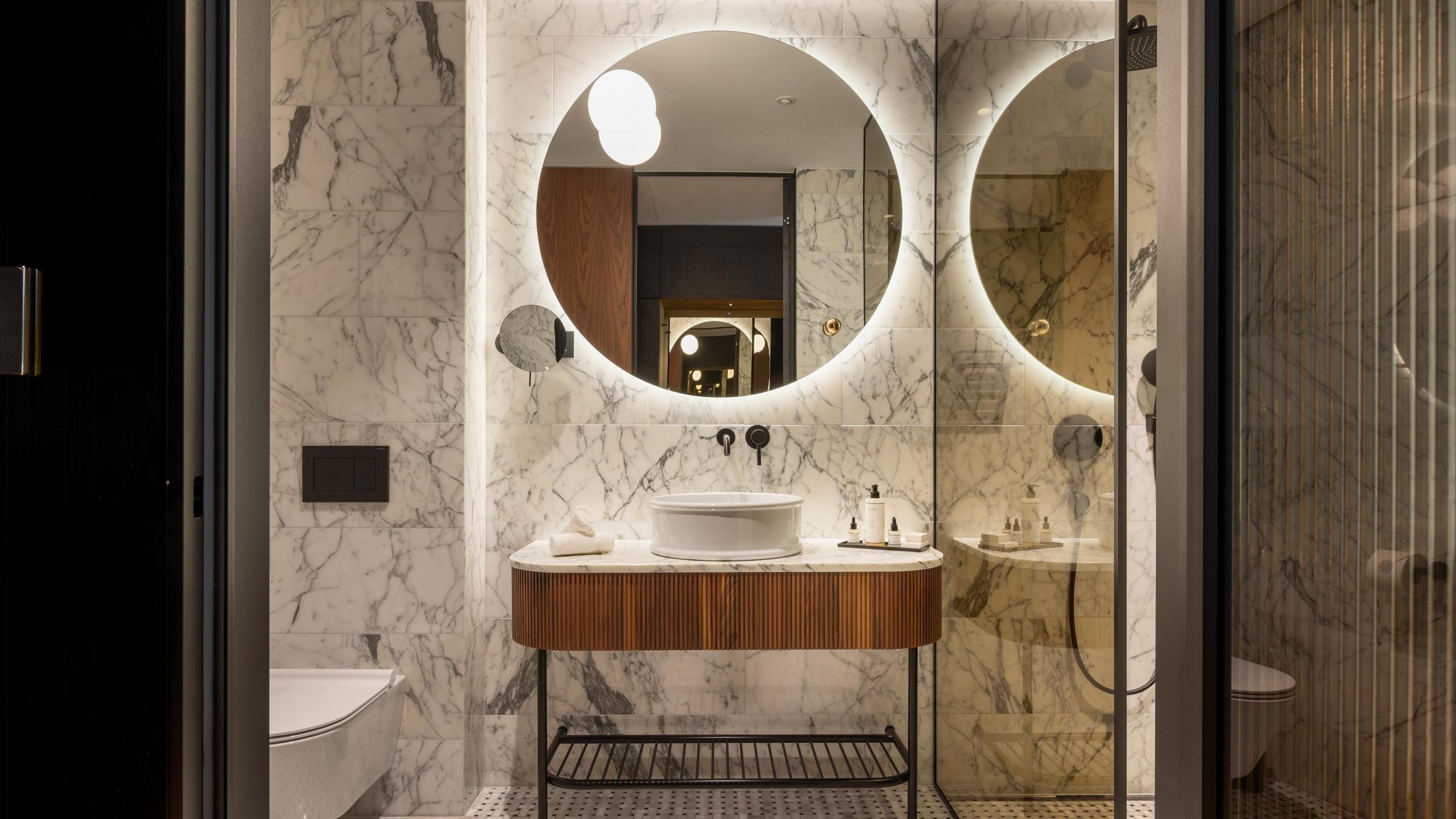 Modern bathroom with a round glowing mirror, marble tiling, and a vessel sink on a fluted wood vanity.
