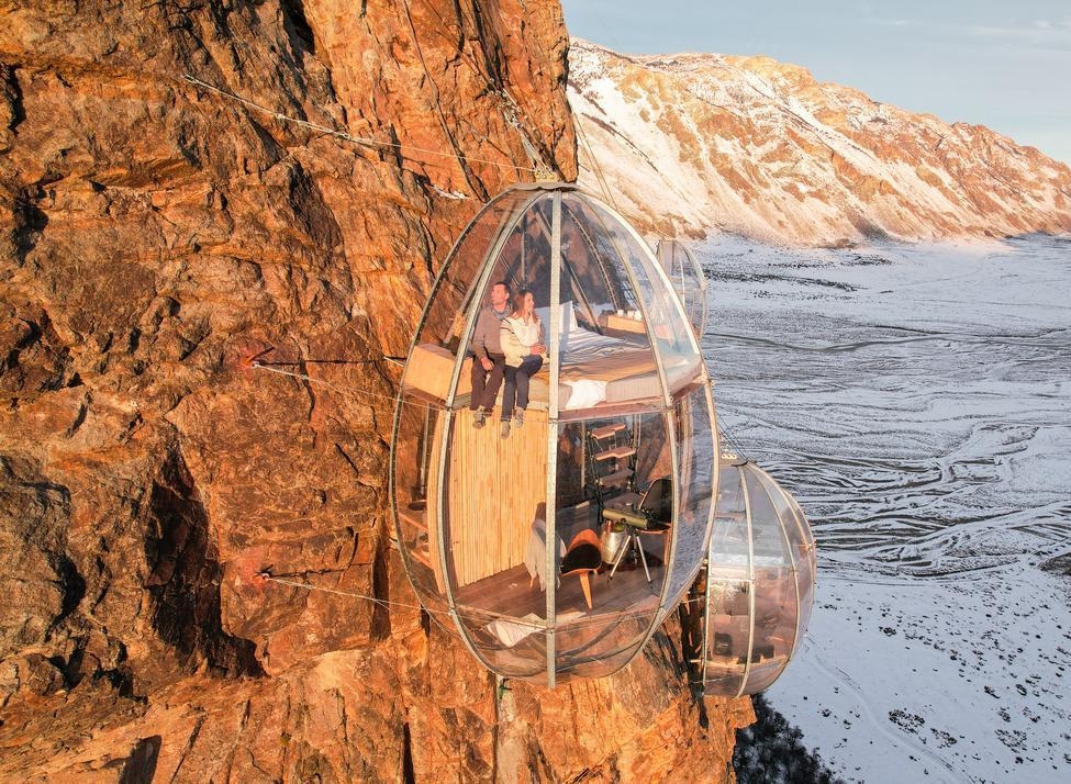 An Ovo Patagonia capsule hanging from a mountain cliff with people inside viewing the winter scenery.