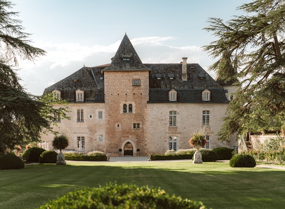 Exterior of a medieval castle hotel with white/beige stone walls and dark tile roof, and a lawn and trees in the foreground.