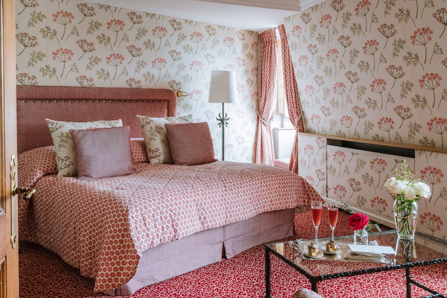 Bedroom interior with a bed with light red bedding, red carpeting and period wallpaper, and a small glass table in the foreground with drinks and flowers.
