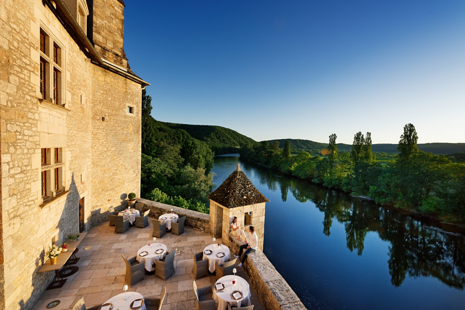 Exterior shot of the castle walls and a terrace with several tables overlooking the Dordogne River and a blue sky.