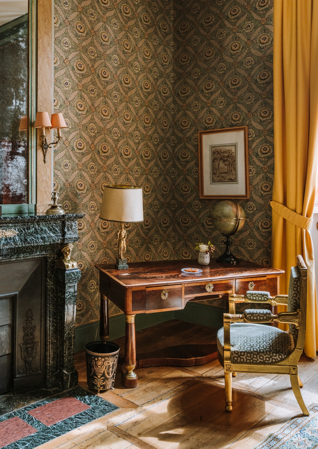Interior shot of a room with antique desk, lamp, chair and fireplace, with a framed picture on the wall, historic wallpaper and a globe on the desk