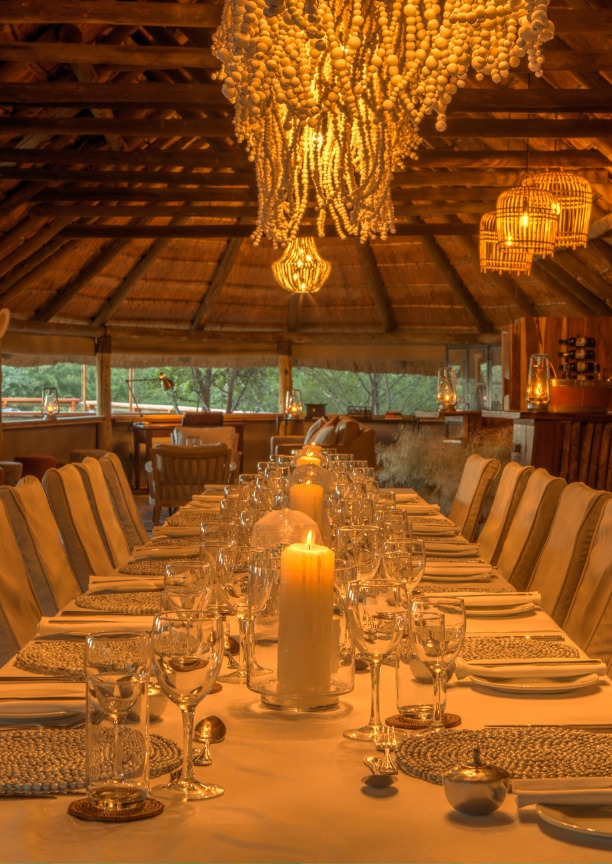 A long formal dinner table set with candles under a thatched roof and beaded chandelier at Dinaka Botswana property.