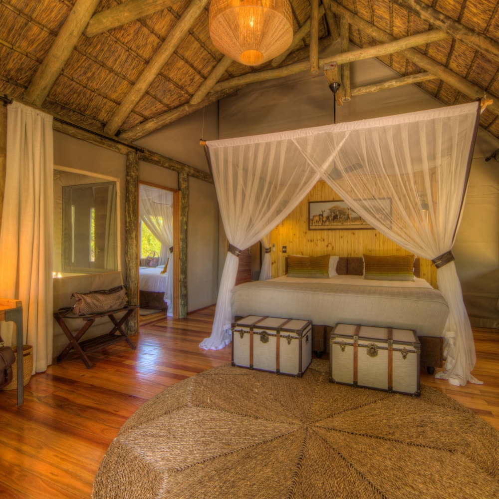 A luxury safari bedroom at Dinaka Botswana property with a mosquito net, thatched roof, and woven rug.