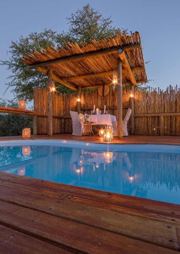 A candlelit dinner table set next to a swimming pool under a wooden structure at Dinaka Botswana property.