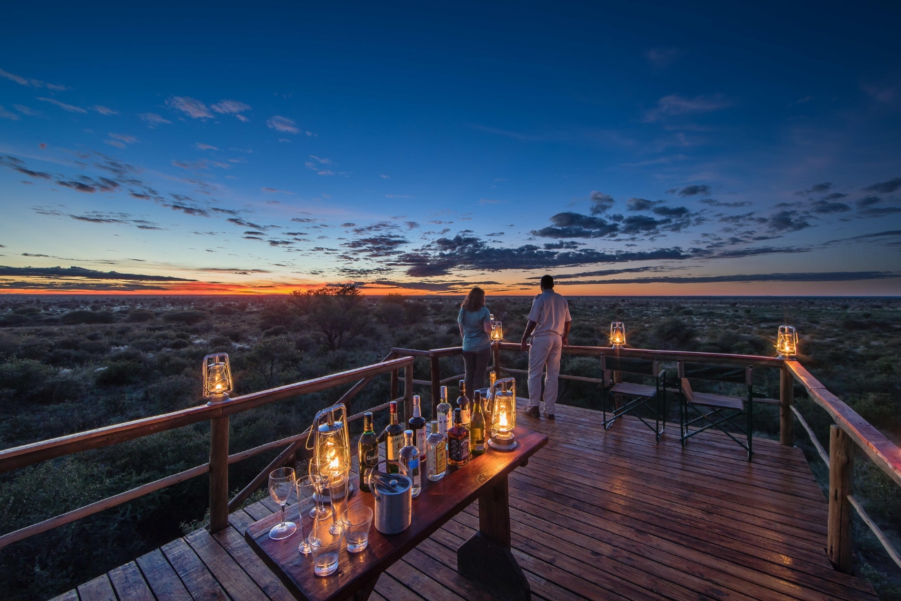 A drinks table with bottles and lanterns on a wooden deck overlooking the sunset at Dinaka Botswana property.