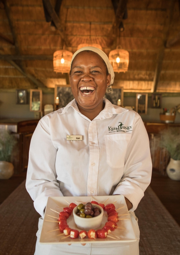 A laughing woman in a white Ker & Downey Botswana uniform holding a plate of tomato and olive snacks.