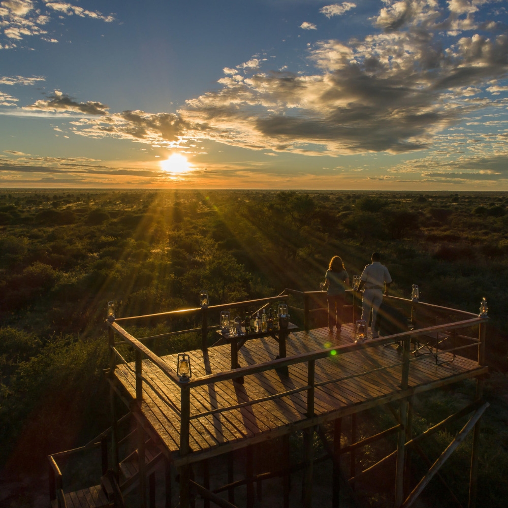 Two people standing on a wooden safari viewing deck at sunset overlooking the Dinaka Botswana property.