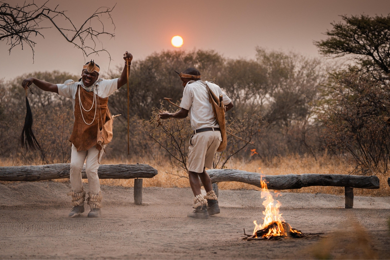 Two men in traditional San attire dancing by a small fire at sunset near the Dinaka Botswana property.