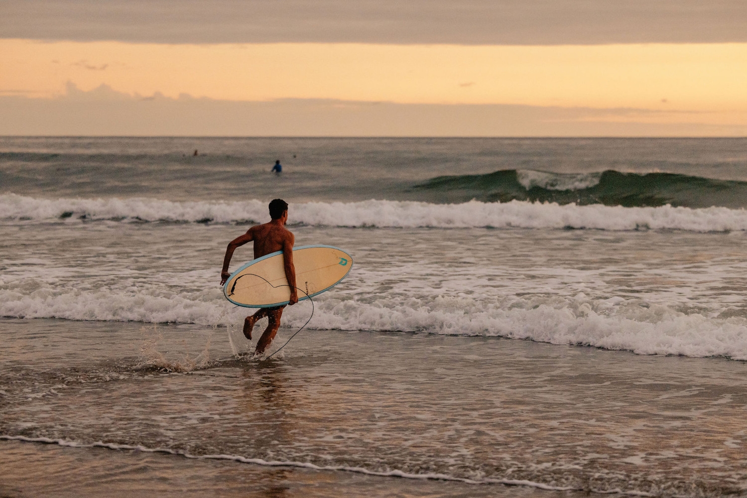 Surfer holding a board running into the waves with sunset in the background.