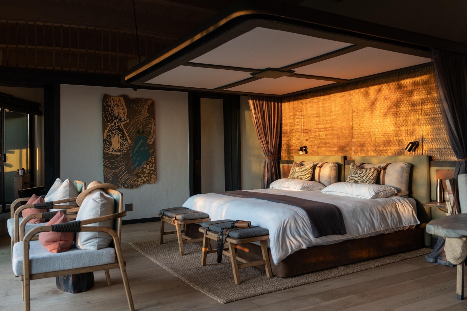 Villa bedroom with a large screen over a double bed with white and brown bedding, several stools and chairs made of wood, and a large painting on the back wall.
