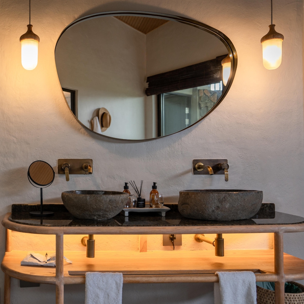Bathroom sink basin with two sinks made of stone and above an irregular-shaped mirror and two hanging bulbs.
