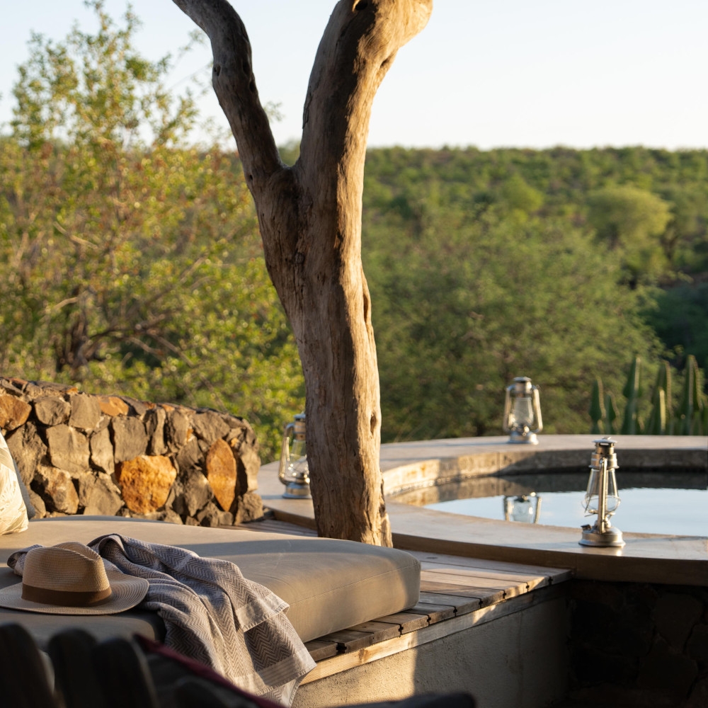 Exterior shot of a lounge chair with a sun hat resting on it. Next to it is a tree trunk and a round plunge pool with the green bushland in the distance.
