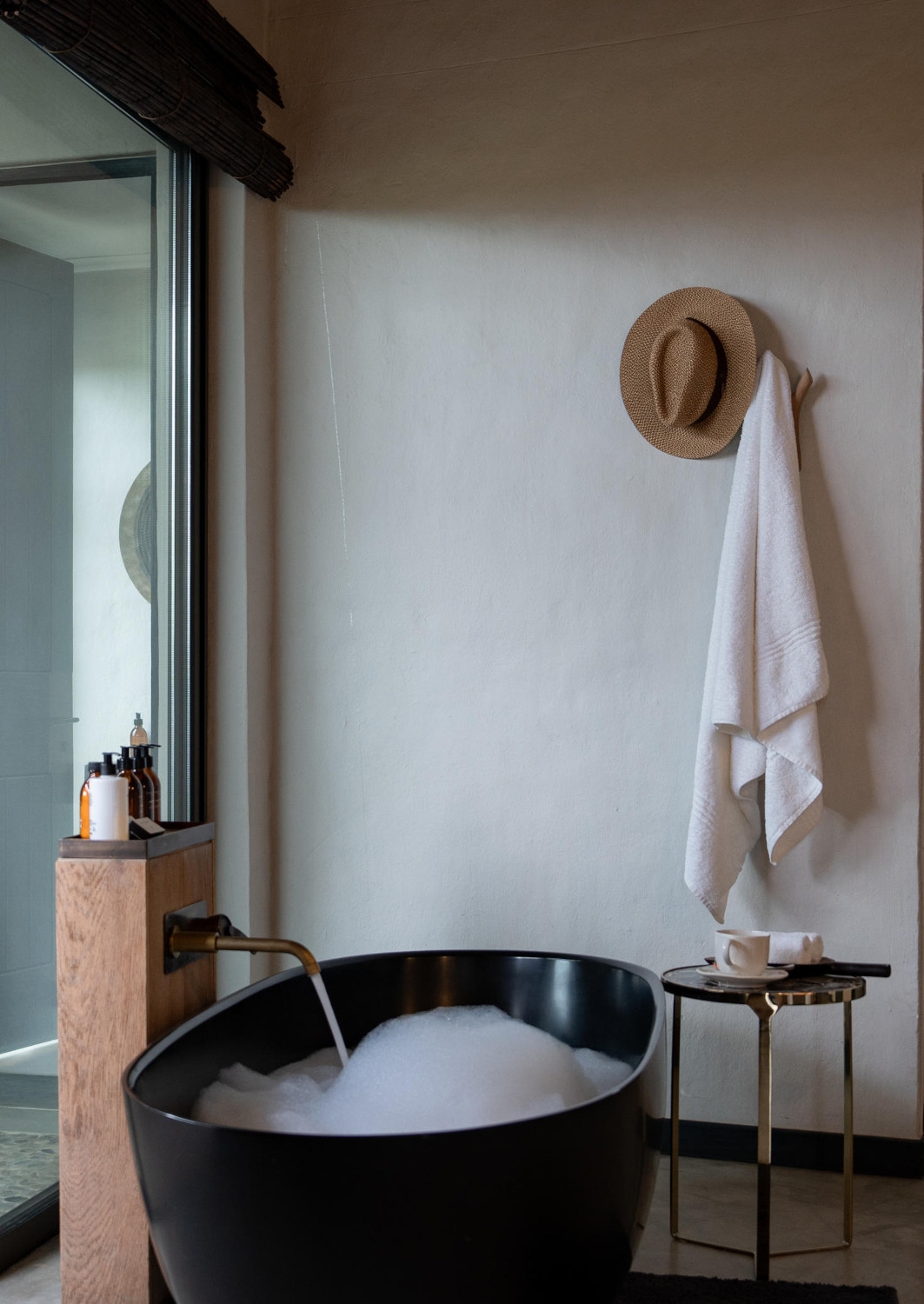 Large black freestanding soaking bathtub mostly full with water and bubbles, and a towel and sun hat hanging on a white wall.