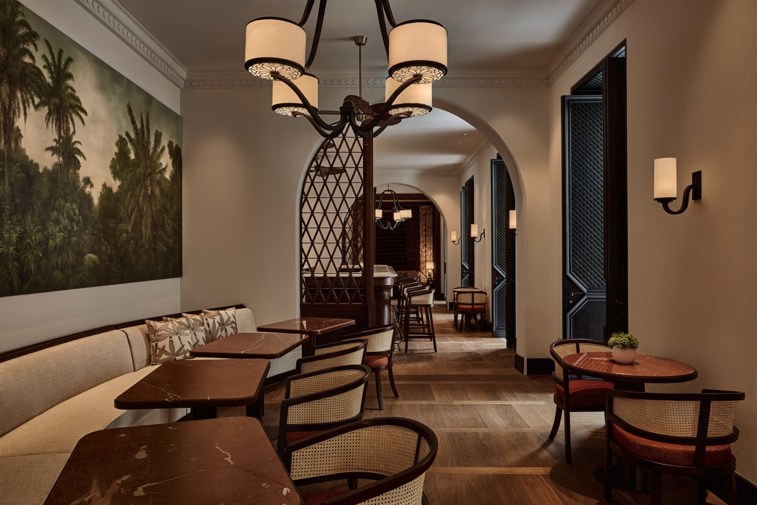 Bar dining area with low beige banquettes and dark marble tables, and midcentury chandeliers. There is an arched doorway into another bar area and a mural on the wall of palms and jungle.
