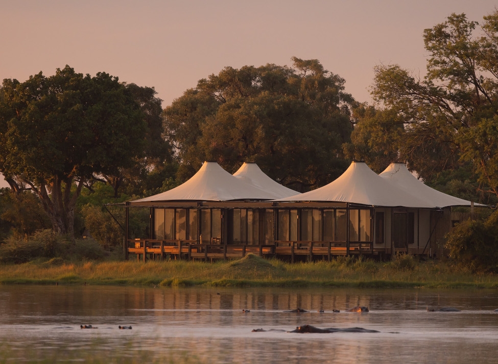 Exterior far-away shot of a tented camp alongside a lake wwith a stand of trees behind it.