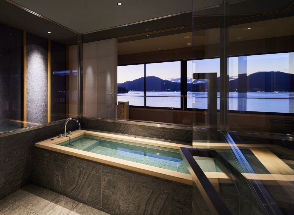 Interior of a corner bath with dual-aspect windows overlooking the sea and mountains with a purple sunset.