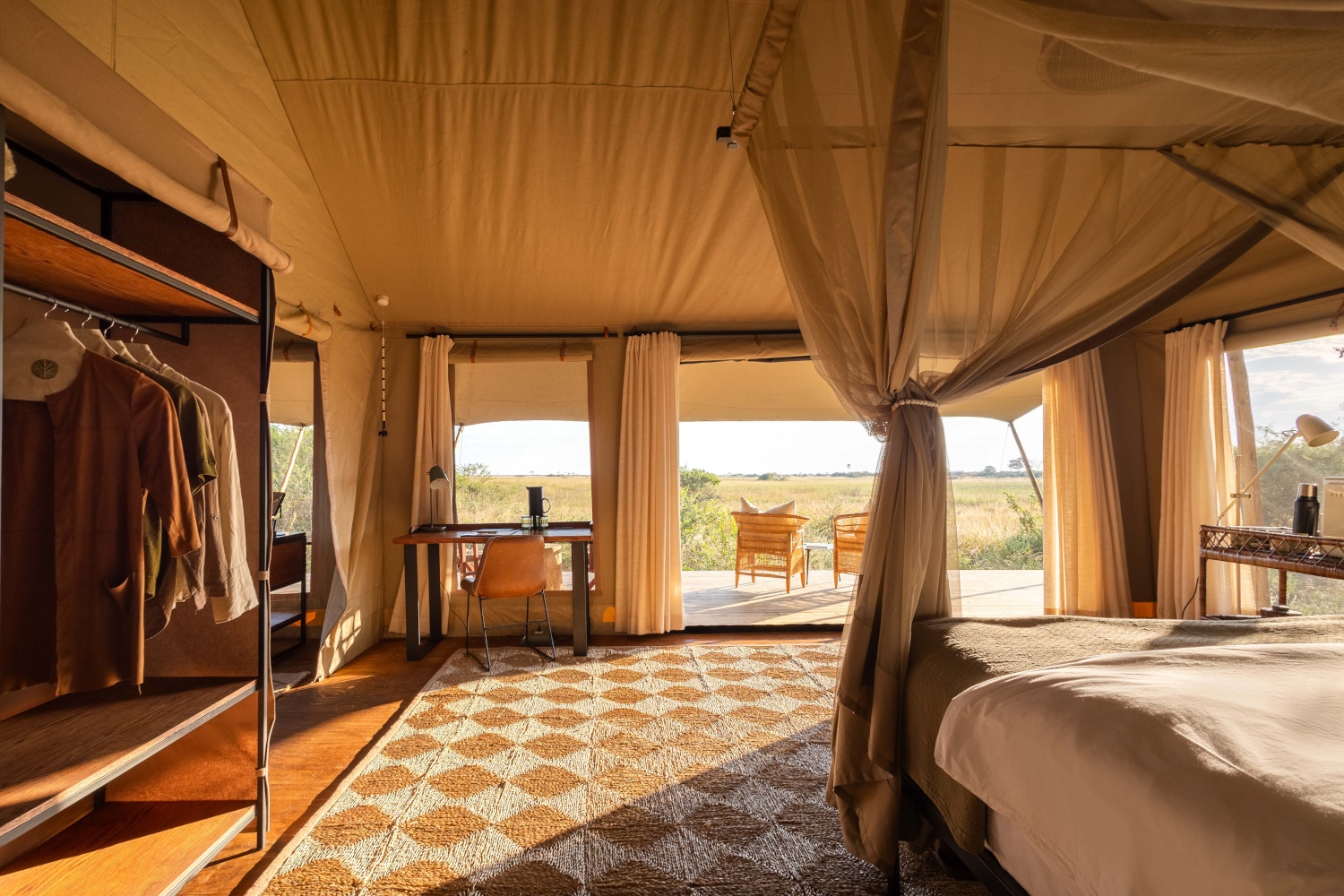 Interior of a safari tent bedroom with a bed on the right and gathered mosquito net and open windows looking into a deck with two chairs and the bush beyond.