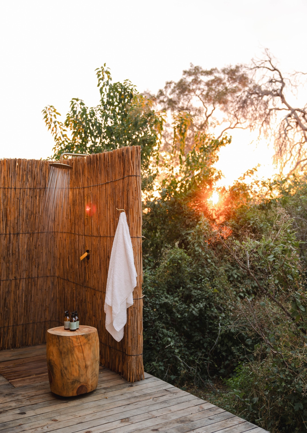 Outdoor shower with a reed fence and greenery just outside and a white towel hanging on a hook.