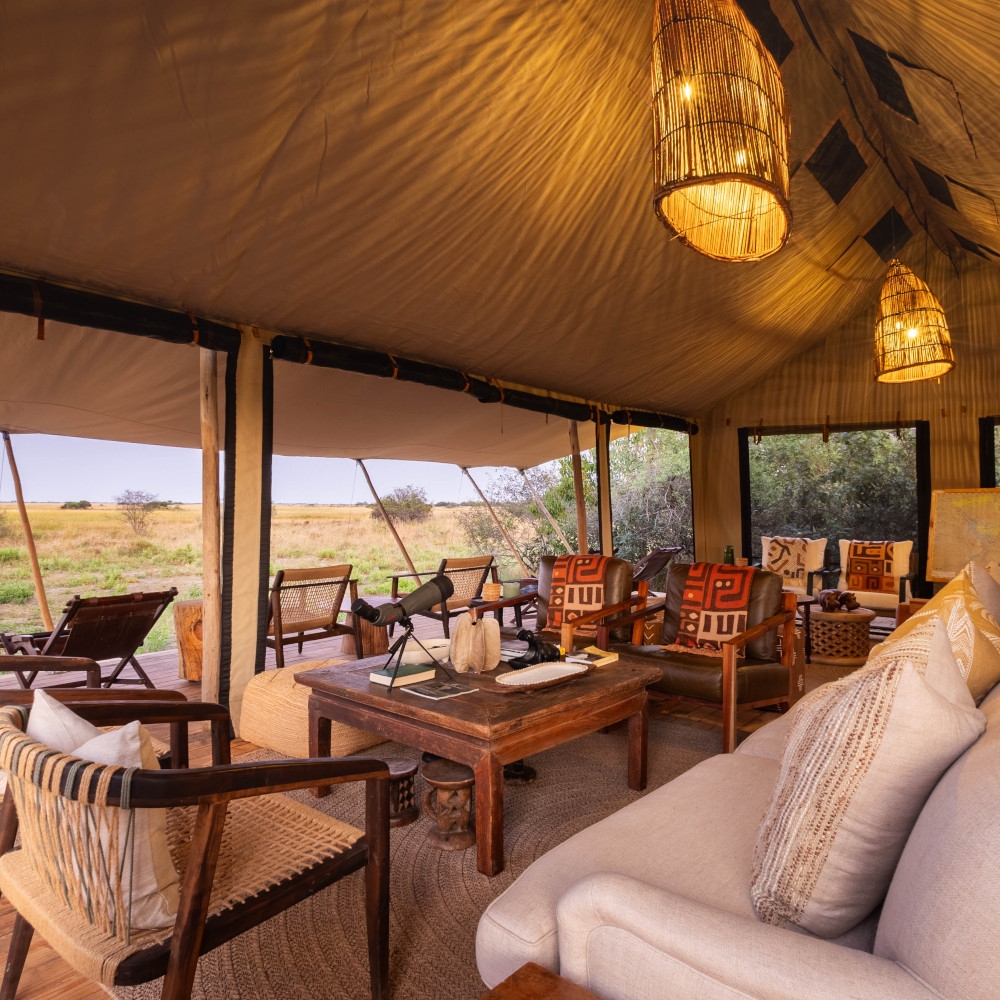 Lounge chairs, dark wood coffee table and a large beige sofa inside a tented canvas lodge area with two lamps overhead and open windows looking across the bush.
