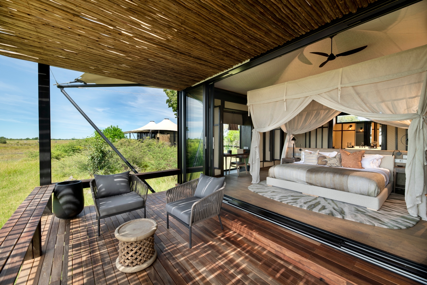 Open-air tented bedroom with a bed with gathered mosquito netting, a wooden floor and wooden ceiling and two lounge chairs looking out over the greenery of the bush and another safari tent in the background.