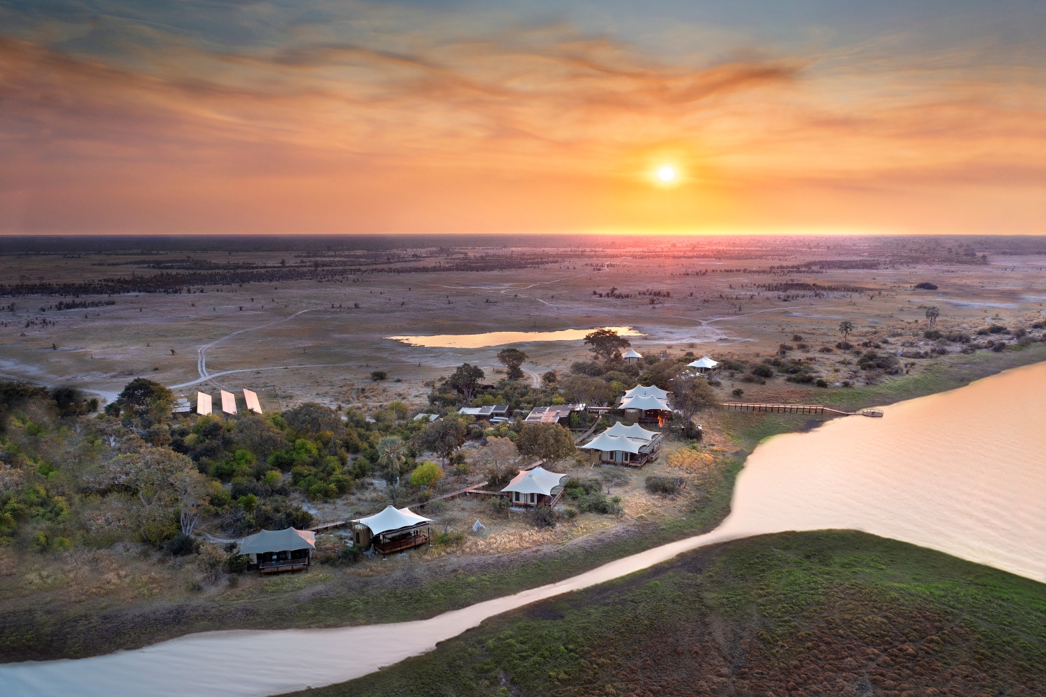 View of a tented safari camp from above with a lagoon flowing along the foreground and the bushland and a yellow sunset beyond.