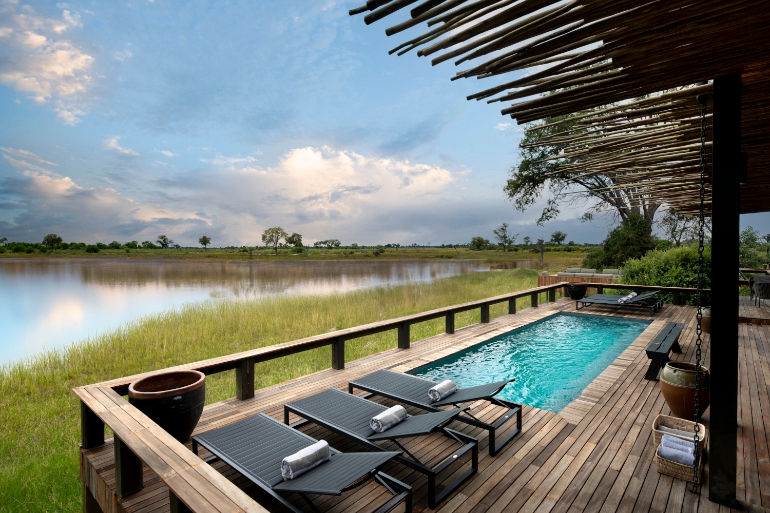 A blue swimming pool with three lounge chairs on a wooden deck and thatch overhand looking out over a wetland with water and grass.