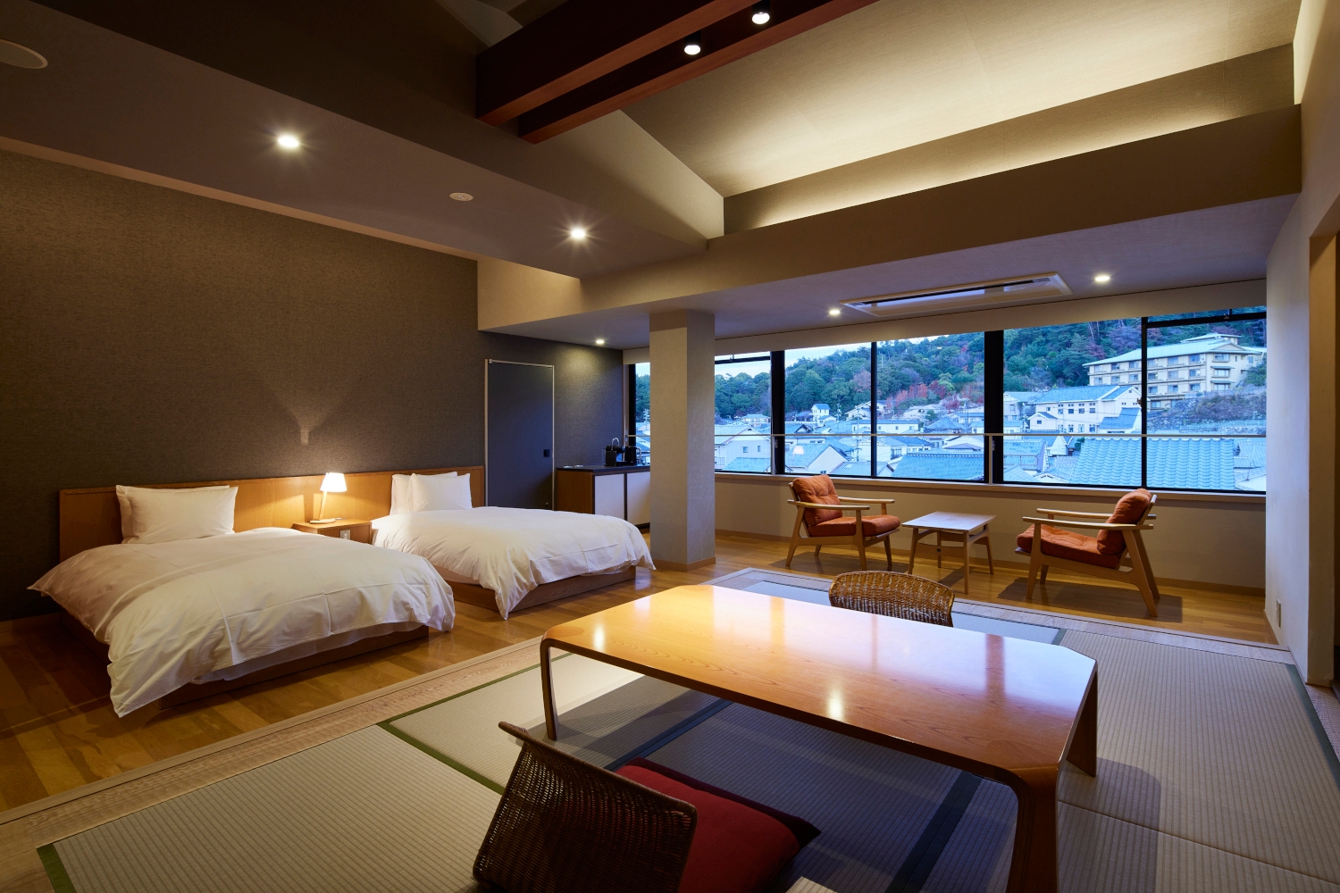 Interior of a double room with two beds with white bedding, a low floor-seating tatami table and a long window looking out over a village and mountain.