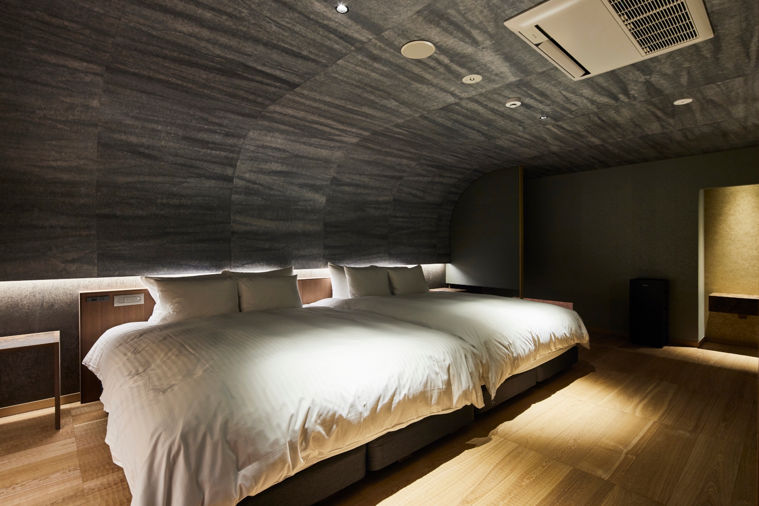 Interior of a double room with two beds with white bedding and rounded stucco ceiling and wood floors.