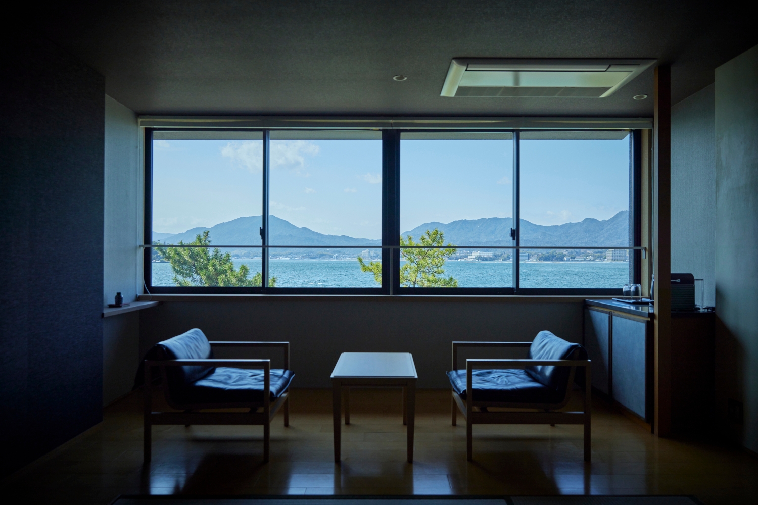 Interior view of a seating area with two vintage chairs facing a small coffee table in front of a long picture window overlooking the sea and mountains.