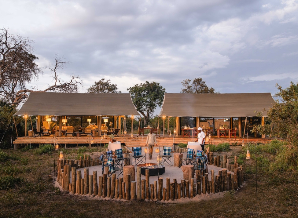 Exterior view of a tented lodge with open walls and a few trees in the background, and people around a fenced firepit in the foreground.