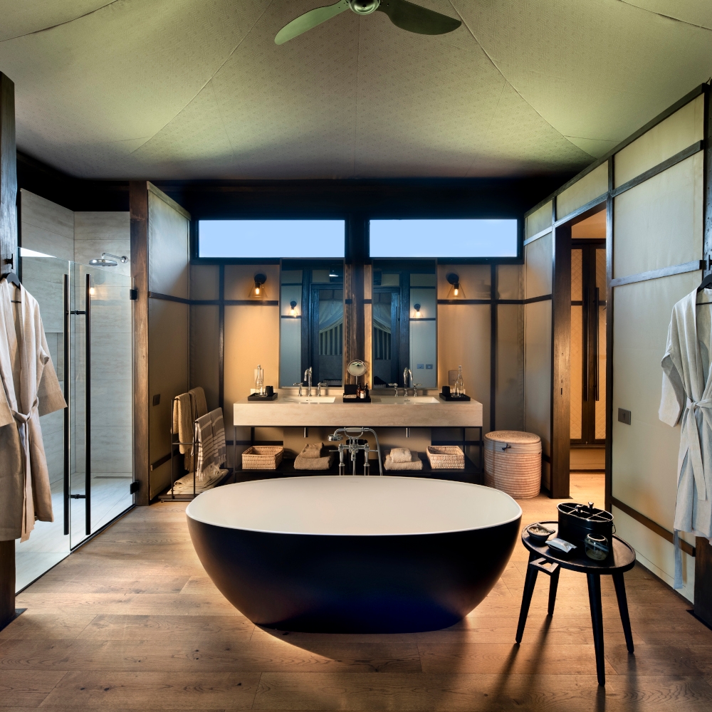 Interior of a safari tent bathroom with a freestanding black tub and small side table, and two bathrobes hanging up and sinks and mirrors in the background.