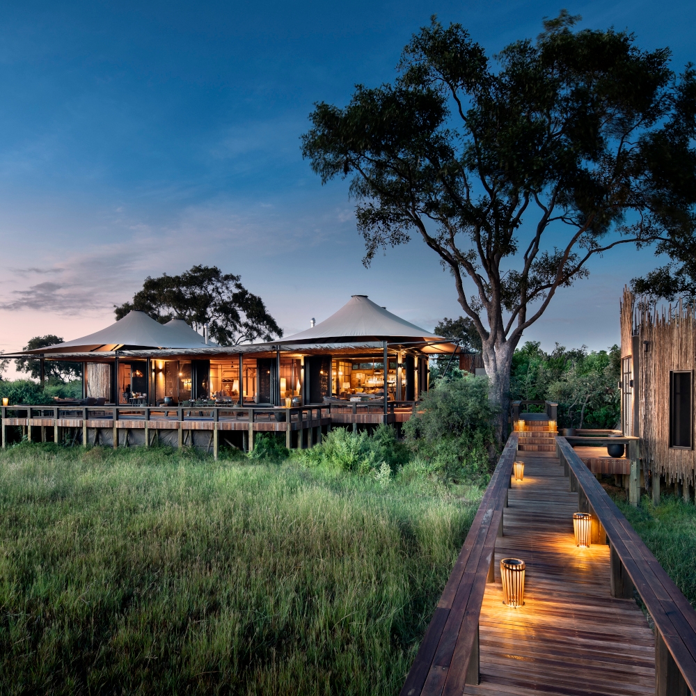 An evening sunset view of a tented safari camp with wooden decking and a wooden walkway lit by small amber lanterns.