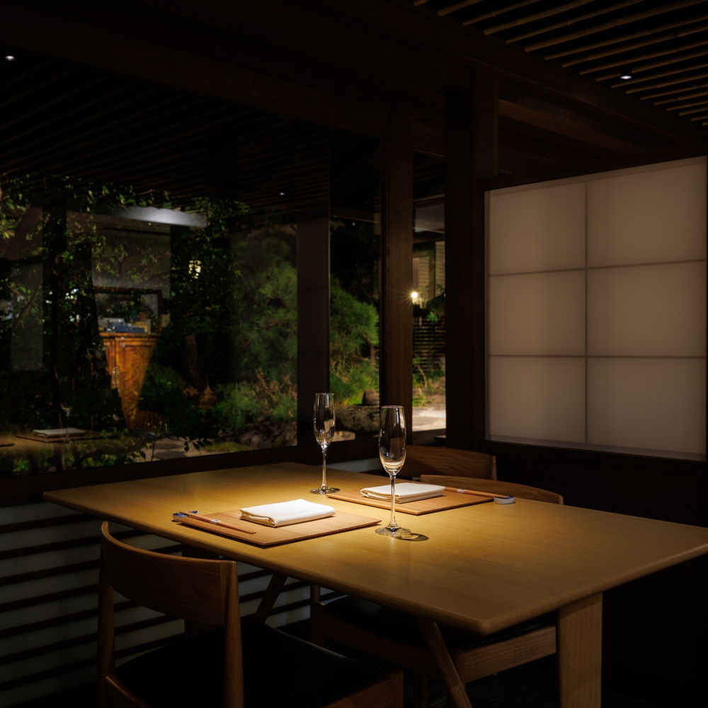 Low-lit dining room with a simple wooden table in the foreground set for dinner, a paper screen in the background and a darkened garden outside a set of windows in the background.