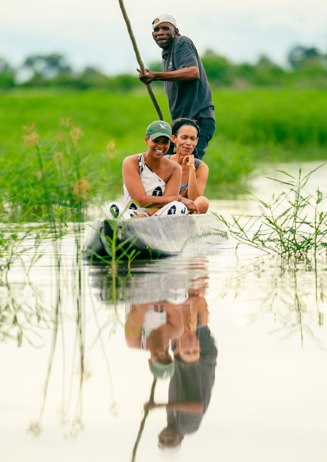 Two people in a small mokoro canoe being paddled by a safari guide at the back, floating through wetland waterway surrounded by green grass.
