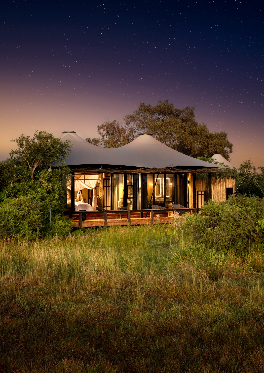 Night view of a safari tent from the outside, with interior lights on and grass in front and trees behind, and a starry sky above.
