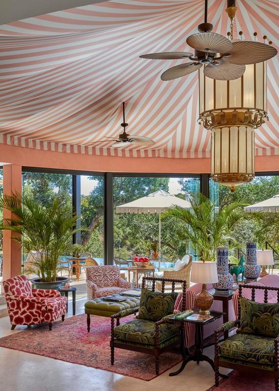 A ceiling fan and glass chandelier hang from a pink and white striped canvas ceiling, above a lounge area with potted palms, patterned armchairs and rugs, with views to the trees outside through tall windows.