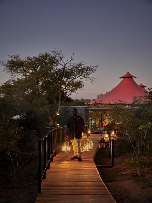 A nighttime scene with a curved wooden walkway leading to red-topped safari tents, with trees at either side. A man in pale trousers and a dark jacket holds several lanterns in each hand.