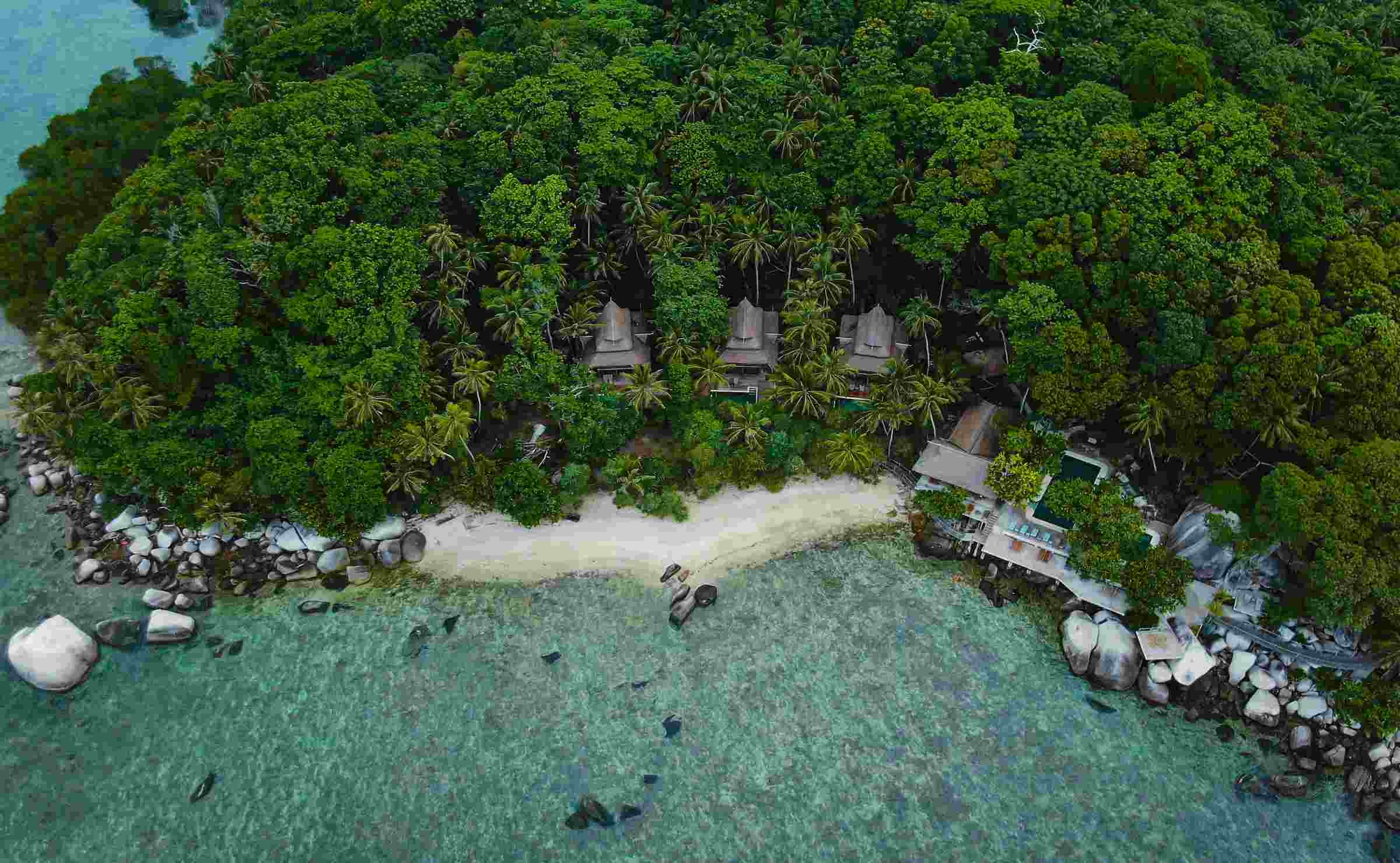 An overhead view of a white-sand beach with dense rainforest and the rooftops of villas visible behind.